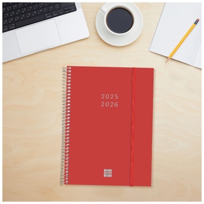 Agenda espiral vermelha 2025 2026 numa mesa junto a portátil, chávena de café e caderno com lápis