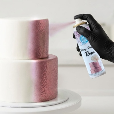 Bolo branco de dois andares sendo pintado com spray metálico rosa por mão com luva preta