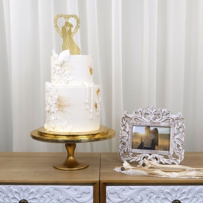 Bolo de casamento decorado branco com suporte dourado e moldura branca com fotografia ao lado