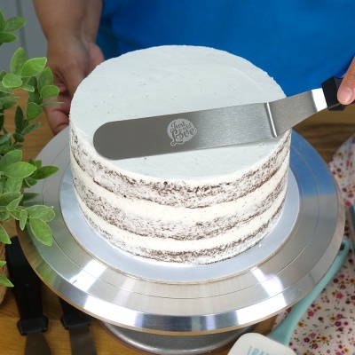 Bolo com cobertura branca e camadas visíveis de bolo de chocolate numa base metálica, com espátula gravada, em ambiente de cozinha.