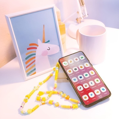 Mesa com moldura de unicórnio, caneca branca, smartphone com ecrã e cordão colorido.