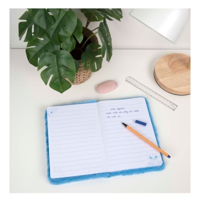 Caderno azul felpudo aberto com caneta e borracha ao lado numa secretária branca com planta