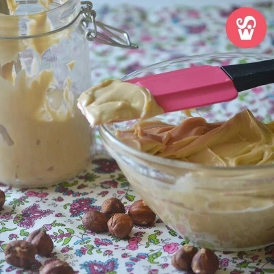 Tigela com creme castanho claro, espátula rosa preta, frasco de vidro e avelãs sobre toalha floral