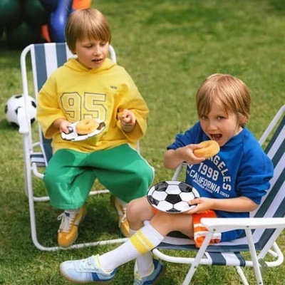 Duas crianças sentadas em cadeiras de praia brancas com riscas azuis, usando roupas coloridas, segurando pratos com padrão de bola de futebol e comendo donuts num relvado verde.
