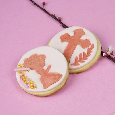 Biscoitos decorados com anjo e cruz em fundo rosa com ramo de flor