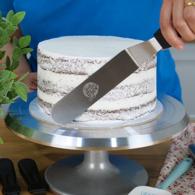 Bolo naked cake branco numa base giratória metálica, com espátula gravada a alisar o bolo.