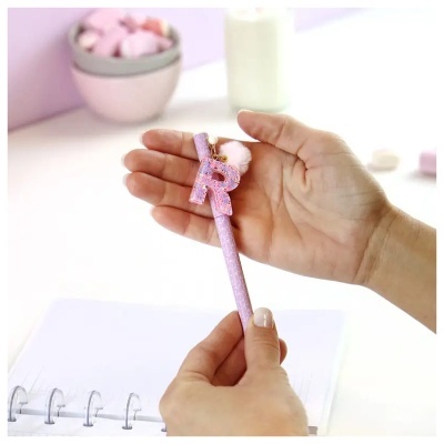 Caneta cor-de-rosa com letra R e pompom cor-de-rosa segurada por mãos sobre caderno