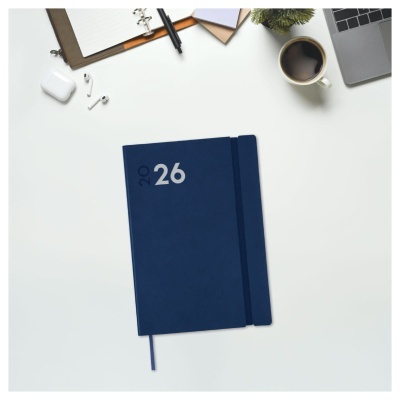 Caderno azul com o ano 2026 na capa sobre mesa branca com vários objetos de escritório