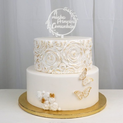Bolo branco decorado com flores e borboletas douradas e topo com texto A minha Primeira Comunhão.
