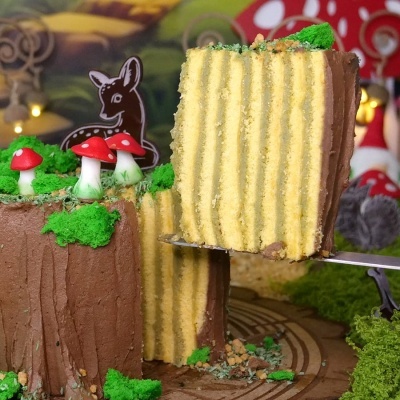 Bolo decorado como tronco de árvore com cogumelos vermelhos e musgo verde, fatia cortada mostrando camadas interiores