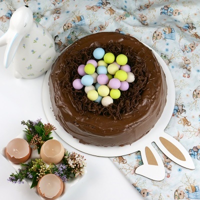 Bolo de chocolate decorado com confeitos coloridos e lascas de chocolate sobre pano com padrão de coelhos, jarra branca e velas em casca de ovo com flores