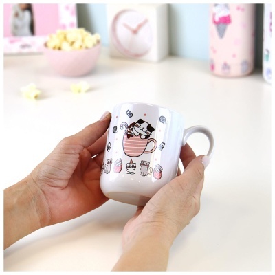 Caneca branca com desenho de gato dentro de chávena rosa, rodeado de doces, segurada por mãos.