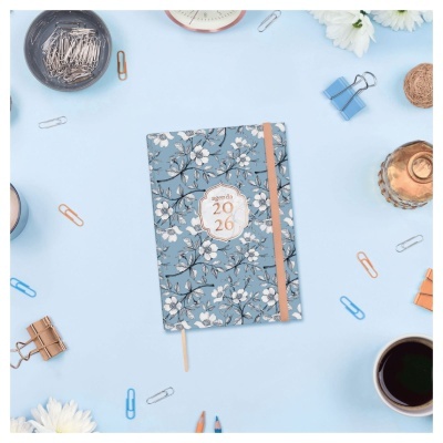 Agenda 2026 com capa azul floral, elástico bege e vários acessórios de escritório à volta sobre fundo azul claro.