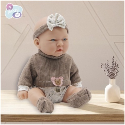 Boneca bebé vestida com roupa castanha e branca sentada numa mesa de madeira ao lado de um vaso branco com ramo seco