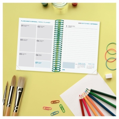Plano semanal 2023 em espiral com pincéis, lápis de cor e clips numa mesa amarela