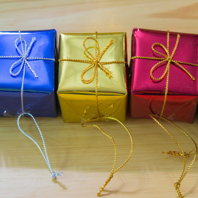 Cinco pequenas caixas de presente coloridas com acabamento metálico e cordão brilhante sobre mesa de madeira.