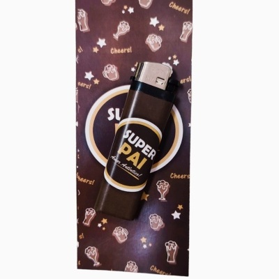 Isqueiro preto com etiqueta SUPER PAI colocado sobre embalagem decorada com canecas e estrelas.