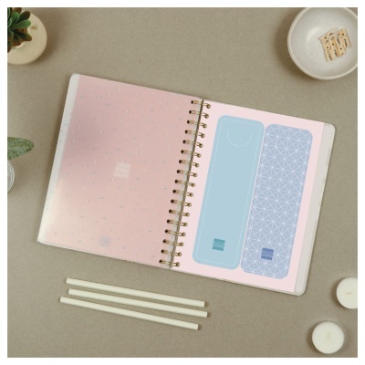 Caderno aberto com marcadores de página azuis em superfície com velas, lápis, plantas e bolachas