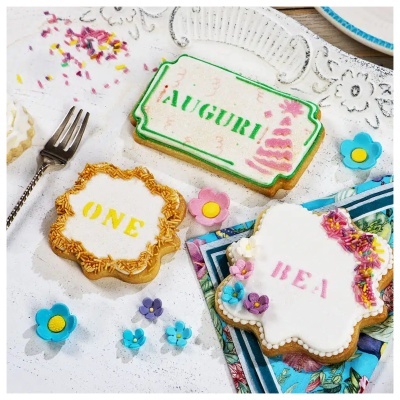 Biscoitos decorados com glacê branco, texto AUGURI!, ONE e REI, e flores de açúcar coloridas