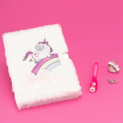 Caderno branco de pelúcia com desenho de unicórnio em arco-íris, caneta rosa e dois pequenos objetos sobre fundo rosa.