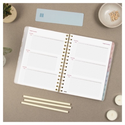 caderno espiralado aberto com páginas quadriculadas e títulos em inglês sobre superfície cinza com lápis, velas, prato com bolachas e plantas