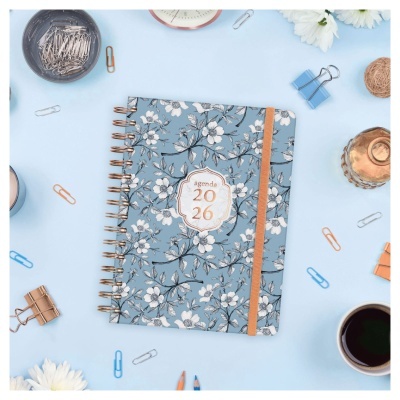Agenda 2026 azul com padrão floral branco e espiral dourada rodeada de clips e objetos de escritório sobre fundo azul