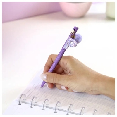 Caneta roxa com pendente P e pompom roxo na mão escrevendo em caderno quadriculado