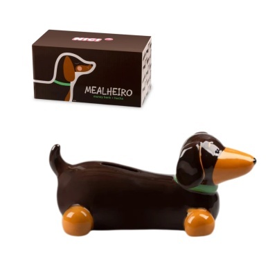 Mealheiro em forma de cão salsicha castanho com detalhes laranja e verde ao lado da sua embalagem.