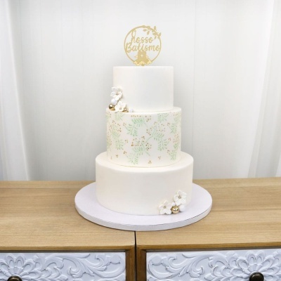 Bolo de três andares decorado com flores e folhas verdes, com topo dourado a dizer Nosso Batismo.
