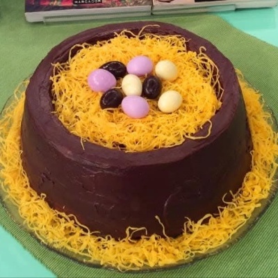 Bolo redondo com cobertura de chocolate e decoração em amarelo, com ovos de chocolate no topo.