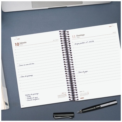 Agenda espiral aberta mostrando páginas de dois dias sobre mesa azul com caneta preta