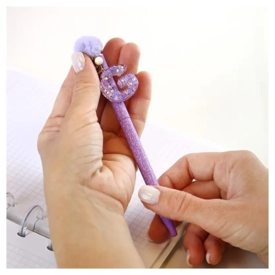 Caneta roxa decorada com letras brilhantes e pompom na mão sobre caderno quadriculado