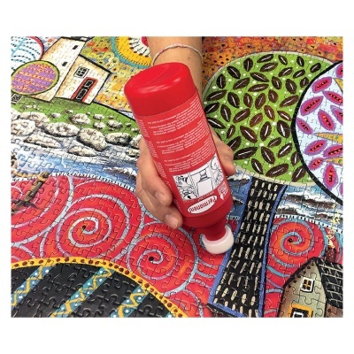 Cola vermelha com aplicador de espuma numa mão sobre puzzle colorido com casas e árvores