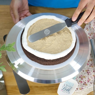 Bolo de chocolate com cobertura bege e borda branca numa base giratória de metal, espalhando creme com espátula