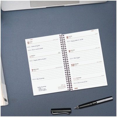 Agenda diária aberta com caneta preta e parte de portátil sobre mesa cinzenta