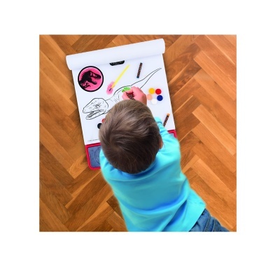 Criança pintando caderno de desenho com ilustrações de dinossauro e materiais coloridos no chão de madeira