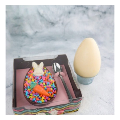 Ovo de chocolate colorido com figura de coelho e cenoura, colher de metal e ovo de chocolate branco em suporte cinzento