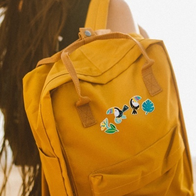 Mochila amarela com autocolantes de tucanos e folhas tropicais.