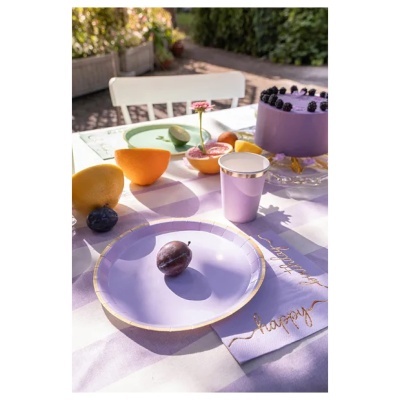 Mesa ao ar livre com pratos e copos lilás, bolo e frutos sobre toalha lilás às riscas.