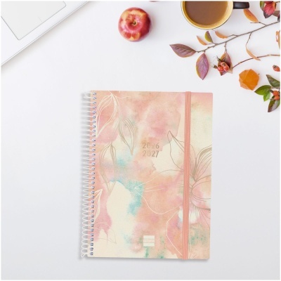 Caderno espiral com padrão aquarelado e elástico rosa, maçã, caneca e folhas sobre mesa branca