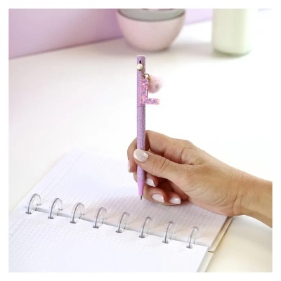 Caneta rosa com chaveiro de pompom numa mão escrevendo num caderno quadriculado