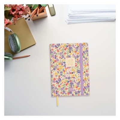 Agenda 2025 2026 floral numa mesa branca com cadernos, lápis, auscultadores verdes e papéis.