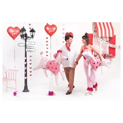 Casal segurando balões em forma de cães rosa com corações, balões de coração vermelhos com texto love you, decoração romântica com candeeiro, coração e janela com toldo.