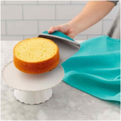 Bolo amarelo redondo sendo retirado com espátula de silicone azul do suporte branco.