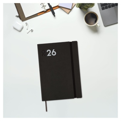Planificador anual preto com número 26 branco na capa sobre mesa com vários acessórios