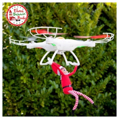 Drone branco com hélices vermelhas segurando boneco de elfo vermelho e riscas