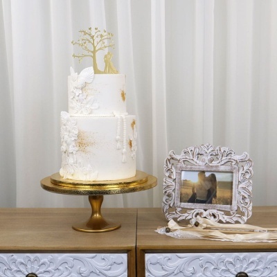 Bolo de casamento branco decorado com flores em relevo e detalhes dourados ao lado de um porta-retratos com foto de casal