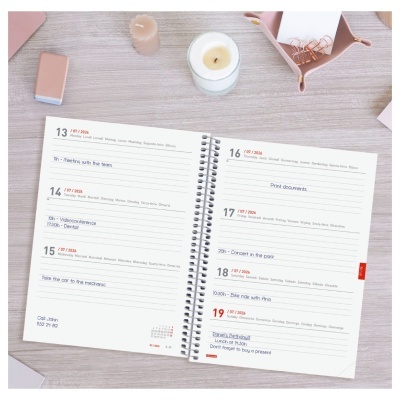 Agenda semanal em espiral com texto impresso numa mesa clara com vela, isqueiro e caixa cor de rosa