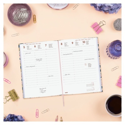 Agenda semanal aberta num fundo bege com clips de papel, flores secas roxas, canetas e copo de café