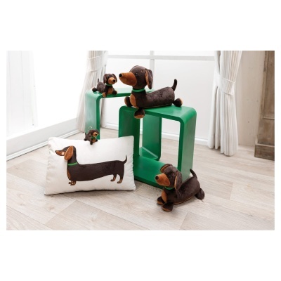 Peluches de cão salchicha castanho e almofada branca com figura bordada, mesas verdes e cortinas brancas no fundo.